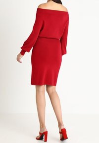 Abito rosso a maglia off-the-shoulder con maniche lunghe, vita aderente e orlo a lunghezza ginocchio. Abbinato a scarpe con tacco alto rosse. Texture liscia.