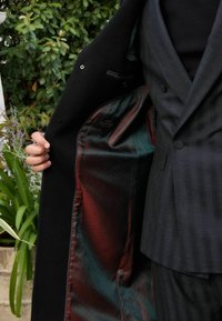 Abrigo negro con un forro de patrón verde profundo y rojo. Presenta un diseño de doble botonadura y una textura suave, con la etiqueta interior visible.