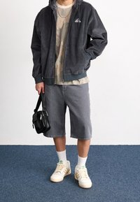 Man som bär en svart Adidas-korsettjacka, kamouflagemönstrad t-shirt, grå shorts, vita Adidas-sneakers och håller en svart crossbody-väska.