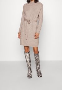 Abito lavorato a maglia beige con collo alto, vita accentuata da una cintura e maniche lunghe, abbinato a stivali al ginocchio neri e bianchi con motivo.