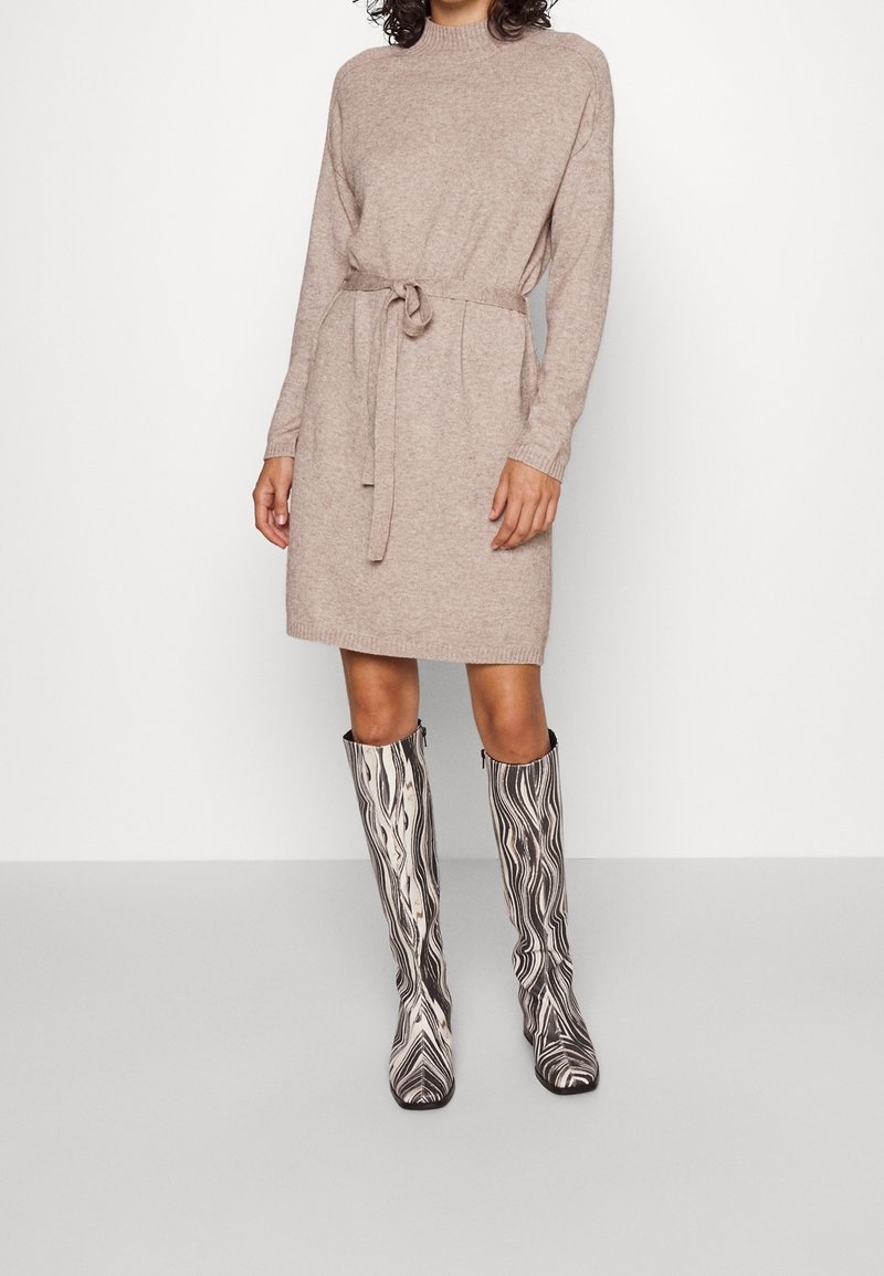 Abito lavorato a maglia beige con collo alto, vita accentuata da una cintura e maniche lunghe, abbinato a stivali al ginocchio neri e bianchi con motivo.