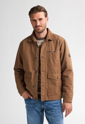 Brune katoenen jas met knoopsluiting, twee borstzakken en een relaxed fit. Gecombineerd met een geribde trui en blauwe jeans.