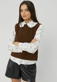 Jeune femme aux longs cheveux bruns, portant un pull sans manches marron par-dessus une blouse blanche à motifs floraux et un short en cuir noir, les bras croisés.