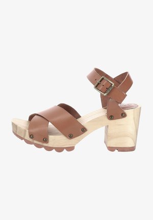 Sandale à talons en cuir marron avec des brides croisées, talon en bois et boucle en métal. Semelle texturée avec des bords festonnés.