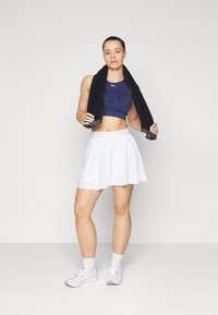 Tengerés sportmelltartó diszkrét mintával, fehér bő szoknya, fekete törölköző a nyakban, és fehér sportcipők. Simító textúrák, sportos fazon.