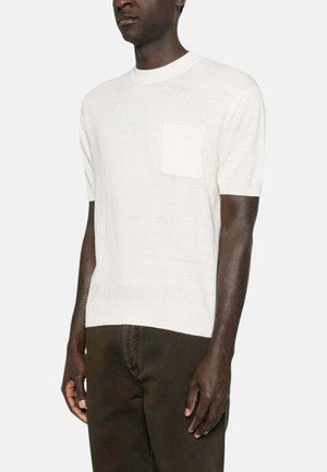 Uomo che indossa un maglione bianco semplice a maniche corte con tasca sul petto e pantaloni scuri su sfondo chiaro.