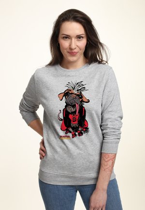 Kvinde med langt brunt hår iført grå sweatshirt med tegneseriehund i Deadpool-kostume og blå jeans, stående mod en lys baggrund.