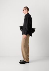 Black button-up shirt, beige wide-leg trousers, black shoes. Model holds a black bag, wearing dark sunglasses. Minimalist background.