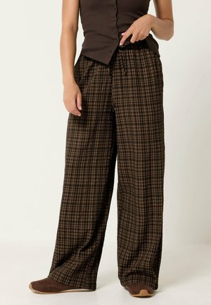 Persona que lleva pantalones sueltos marrones a cuadros, una blusa sin mangas marrón oscuro con botones y zapatos marrones, de pie frente a un fondo liso.