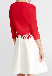 Rød bluse med trekvartærmer og blomster broderi ved taljen, parret med en hvid, vida nederdel. Glat tekstur, tætsiddende overdel.