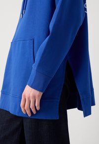 Hoodie bleu à manches longues avec poche avant, fentes latérales, porté avec un pantalon foncé, main détendue le long du corps sur un fond uni.