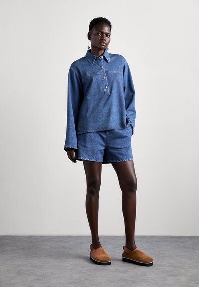 Denim long-sleeve top with a collar and button detail paired with denim shorts, both in a medium blue shade. Brown suede slip-on shoes.