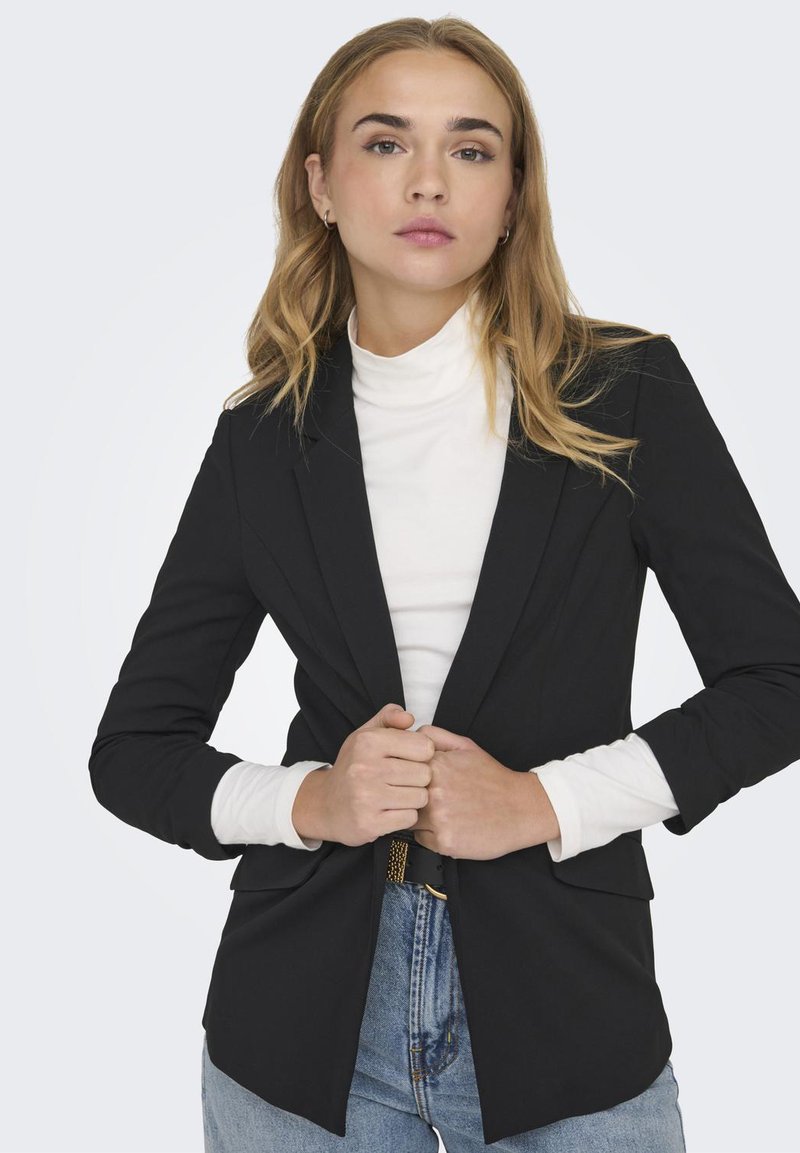 Zwarte op maat gemaakte blazer met een slim fit, voorzien van gestructureerde schouders, uitgeknipte revers en een soepele textuur over een witte coltrui.