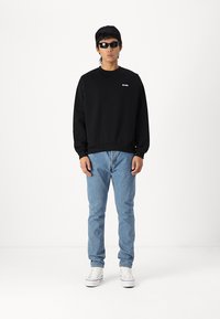 Svart sweatshirt med ribbede mansjetter, parret med lyseblå jeans og hvite sneakers. Modellen har på seg svarte solbriller og står foran en hvit bakgrunn.