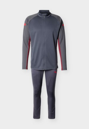 Tmavosivá bunda na zips s červenými pruhmi po bokoch a zodpovedajúce nohavice, oboje s malými červenými logami Puma na rukávoch a stehne.