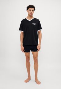 T-shirt preta de mangas curtas com pormenores brancos e pequeno logo, combinada com calções escuros. Design simples, tecido de algodão, corte descontraído.