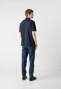 Camisa polo azul-marinho com colarinho, mangas curtas e corte reto, combinada com jeans de ganga azuis e sapatos pretos, vista pela parte de trás.