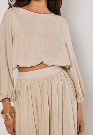 Femme portant un crop top beige texturé à manches longues et une jupe plissée fluide assortie avec des bracelets dorés au poignet gauche.