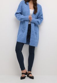 Světle modrý pletený cardigan s kapsami, otevřeným předním dílem, žebrovaným lemem a dlouhými rukávy, nošený přes bílé tričko a tmavé džíny.