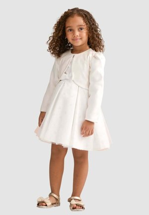 Ragazza con capelli ricci che indossa un vestito bianco, una giacca bolero abbinata e sandali beige con fiocchi, in piedi contro uno sfondo semplice.