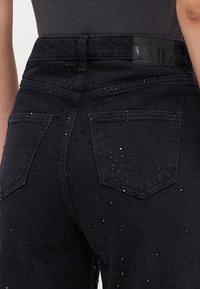 Black denim jeans featuring a fitted design, embellished with small silver and black studs, and a label on the waistband. Classic five-pocket style.