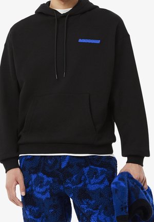 Czarna bluza z kapturem z niebieskim logo Lacoste na piersi, noszona z niebiesko-czarnymi, wzorzystymi spodniami z polaru, trzymająca w prawej ręce pasujący polarowy element odzieży.