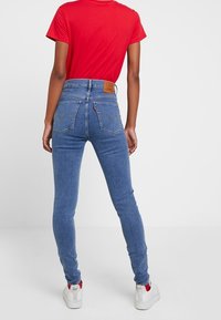 Blue high-waisted jeans with a textured fabric, two back pockets, and a brown leather patch at the waist, paired with a red T-shirt.