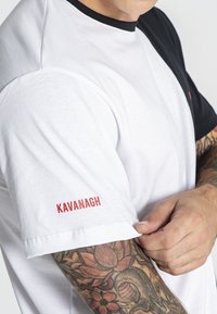 Tričko s krátkym rukávom s bielym telom a čiernymi ramenami, červený text "KAVANAGH" na rukáve, hladká bavlnená tkanina a voľný strih.