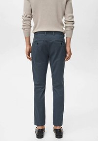 Tmavě modré zúžené kalhoty z bavlněné směsi; mají zadní kapsy na knoflíky a hladký povrch. Nošeny s lehkým svetrem.