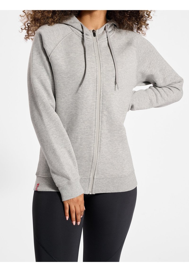 Personne portant un sweat à capuche zippé gris clair avec cordons et manches longues, associée à un legging noir, debout avec un bras plié.