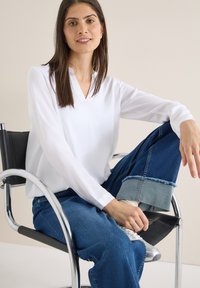 Femme aux longs cheveux bruns, portant un chemisier blanc et un jean bleu retroussé, assise sur une chaise noire et chromée, tenant une basket argentée.