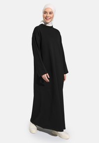 Long black dress made from soft fabric, featuring long sleeves and a loose cut. Worn with white sneakers and a light-colored hijab.