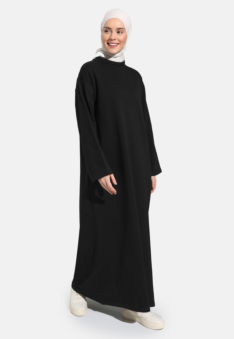 Long black dress made from soft fabric, featuring long sleeves and a loose cut. Worn with white sneakers and a light-colored hijab.