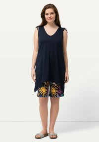 Tunique senza maniche navy con scollo a V e spalline con nodo, abbinata a pantaloni corti colorati con disegni di sole e motivi floreali, indossata con sandali grigi a scivolo.