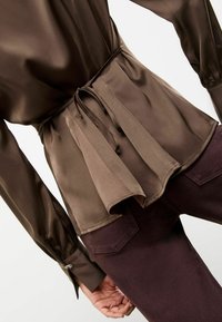 Bluză din satin maronie cu talie ajustabilă, margine plisată și mâneci lungi. Asortată cu pantaloni de culoare purpurie închisă, cu buzunare aplicate.