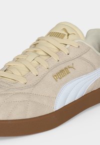 Μπεζ σουέντ αθλητικό παπούτσι Puma με λευκή πλευρική ρίγα, κρεμ κορδόνια, χρυσό λογότυπο Puma στη γλώσσα και στην πλευρά, και καφέ λαστιχένια σόλα.