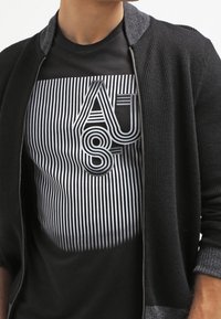 Černá zipová bunda s texturovanou tkaninou a šedými akcenty, vrstvená přes černé tričko s výraznými bílými pruhy a grafickým designem.