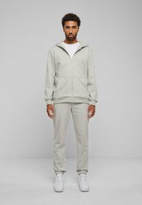 Lys grå zip-up hættetrøje med en frontlomme, parret med matchende joggingbukser og hvide sneakers. Glat stof med en afslappet pasform.
