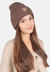 Braune Strickmütze mit geripptem Design und einem goldenen Glitzerherz-Patch. Weicher Stoff, enganliegende Form, getragen mit einem beige gestrickten Pullover.