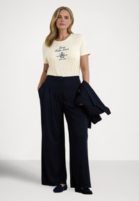 Μπλούζα σε κρεμ απόχρωση με μαύρο λογότυπο, σε συνδυασμό με παντελόνι σε ναυτικό μπλε χρώμα, με φαρδιά πόδια και ψηλή μέση. Κρατώντας σακάκι σε ταιριαστό ναυτικό μπλε. Κλασικά loafers με υφή.