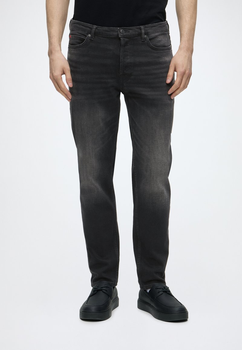 Mand iført slidte sorte slim-fit jeans og sorte snøresko, stående mod en enkel hvid baggrund.