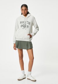 Sudadera gris claro con bolsillo delantero y texto gráfico negro, combinada con una falda plisada verde y zapatillas blancas.