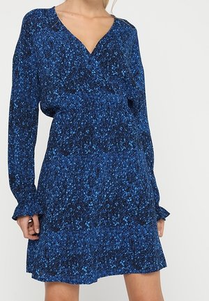 Kobieta ubrana w długą sukienkę z długim rękawem, o wzorze niebiesko-czarnym, z dekoltem w kształcie litery V, z talią podkreśloną i falbankami na rękawach.