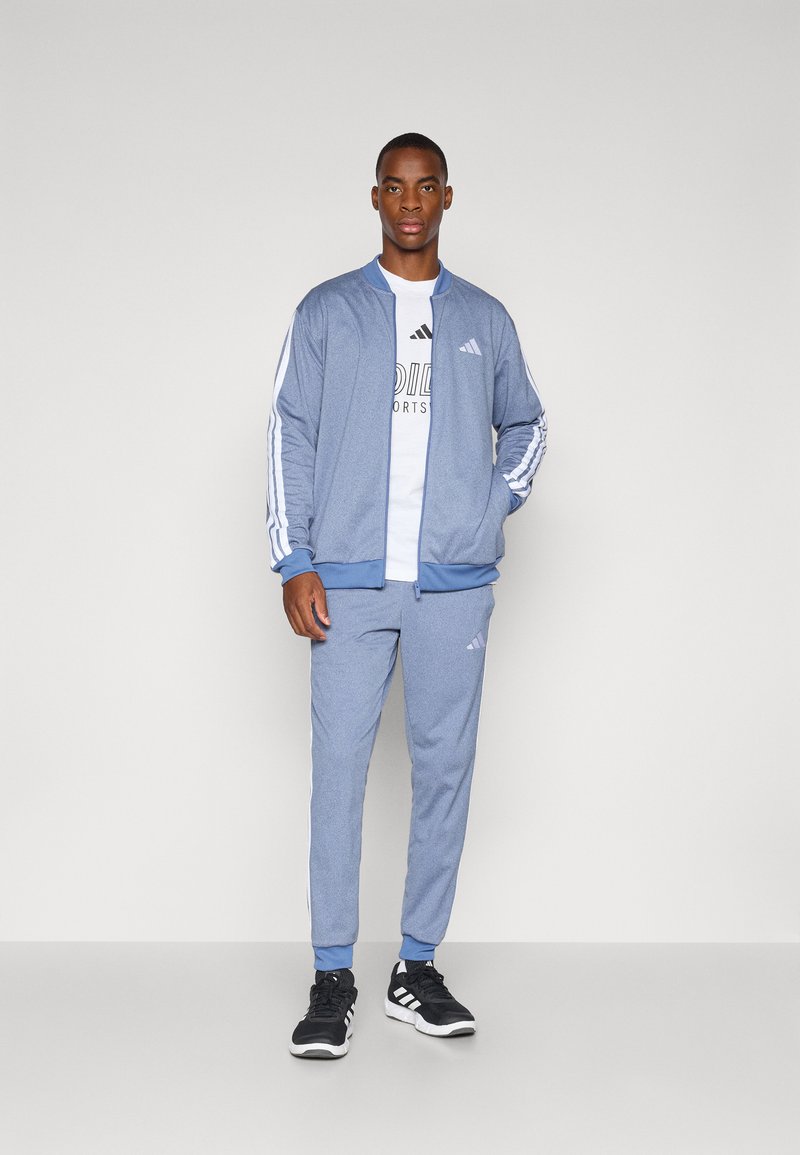 Ensemble de survêtement bleu avec des rayures blanches, veste zippée et poignets élastiques ; associé à un T-shirt blanc et des chaussures de sport noires.