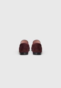 Um par de mocassins em camurça burgundy escuro com solas pretas, vistos de trás sobre um fundo branco liso.