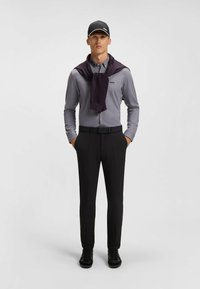 Camicia a quadri grigia, pantaloni scuri, sneakers nere e un maglione viola drappeggiato attorno al collo; berretto nero con logo sulla parte anteriore.