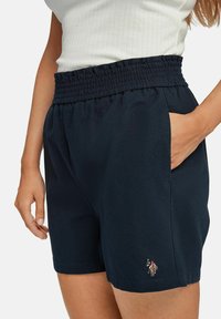 Žena v námořnických modrých elastických šortkách s bočními kapsami a malým vyšitým logem poblíž lemu, v kombinaci s bílým žebrovaným topem.