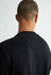 Felpa nera con scollatura rotonda, cuciture visibili e una texture liscia. Il retro presenta un dettaglio di cucitura nella parte superiore.