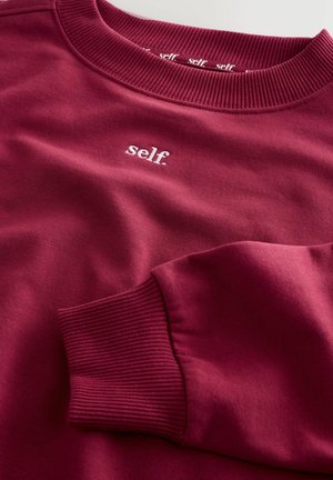 Burgundowa bluza z okrągłym dekoltem i ściągaczami. Z przodu ma małe białe logo "self." na klatce piersiowej. Wykonana z miękkiego materiału bawełnianego w składzie mieszanym.
