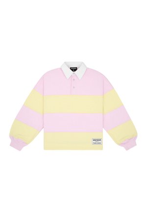 Femme portant un polo à manches longues rayé rose pastel et jaune, un short bleu à carreaux, des chaussettes blanches et des baskets noires, debout devant un fond gris.